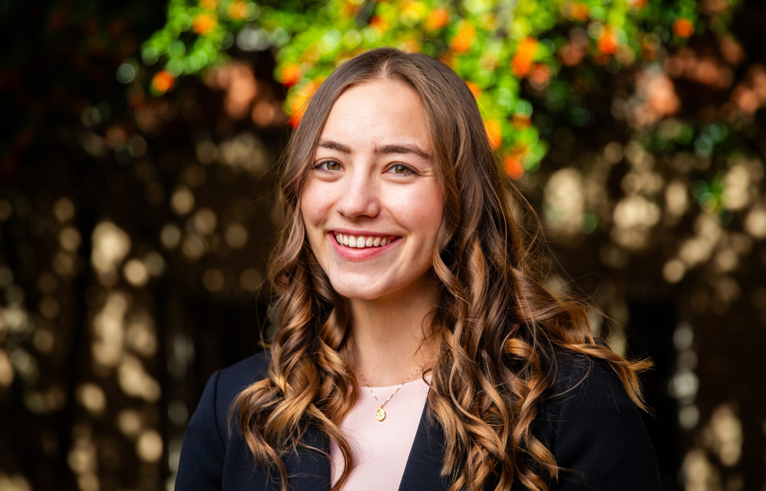 A white woman with brown curly hair smiles in front of fall leaves.