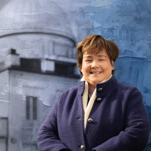 A white woman with short brown hair and a bright blue buttoned jacket stands smiling in front of a graphical image of an observatory behind her.