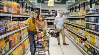 A mother pushes her daughter in a grocery cart down an aisle in the grocery store.