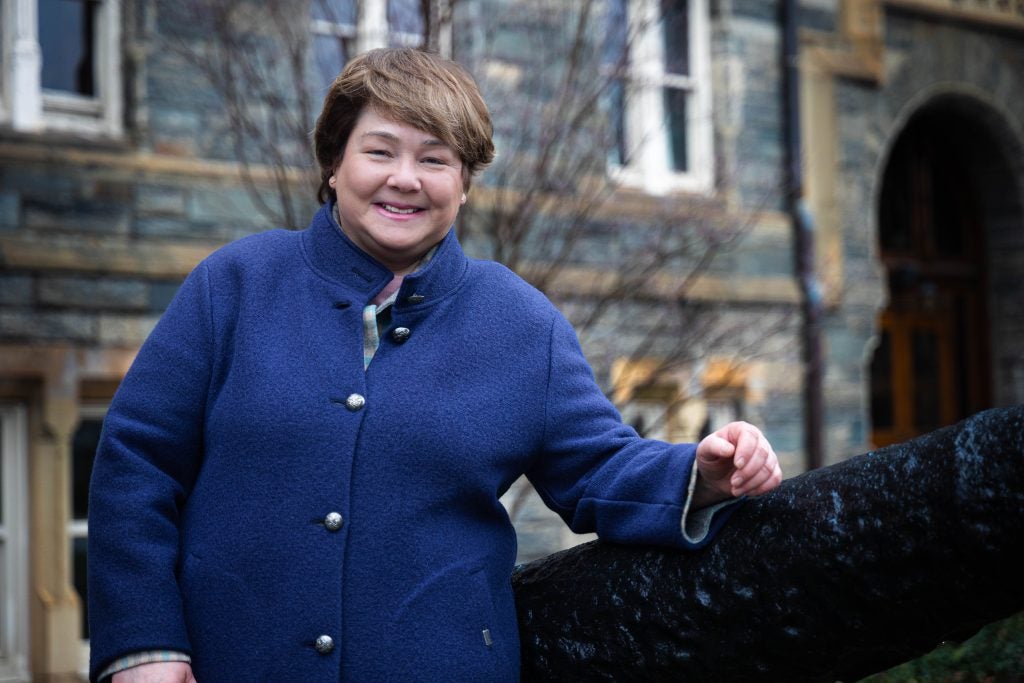 A woman with brown hair and a bright blue jacket leans on a cannon with a stone building behind her.