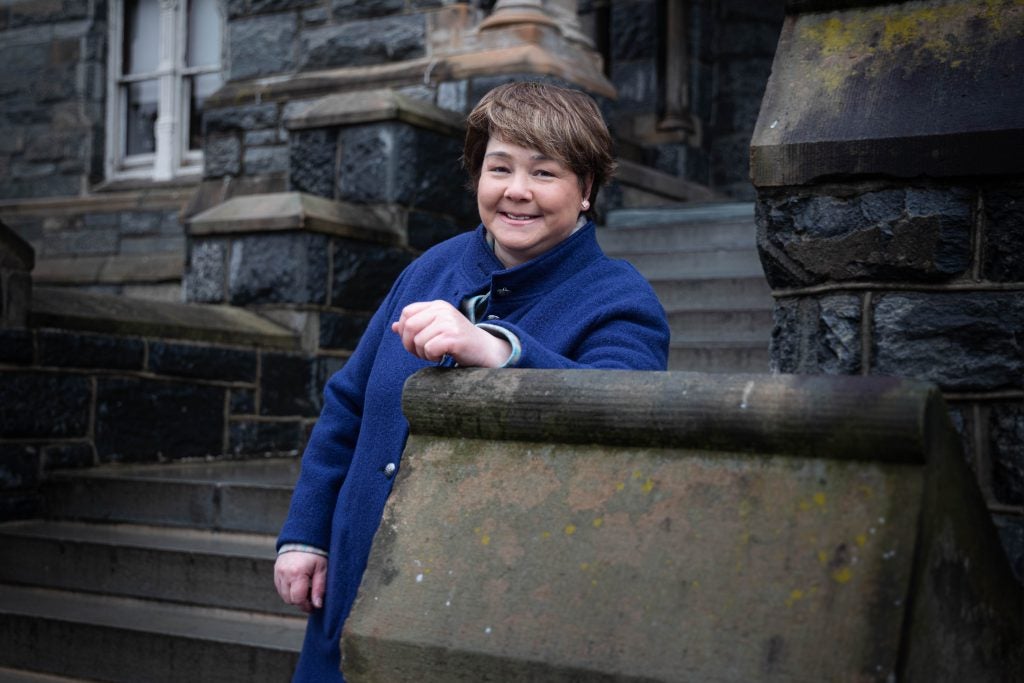 A white woman with brown hair wearing a bright blue sweater leans on a stone ledge with steps behind her.