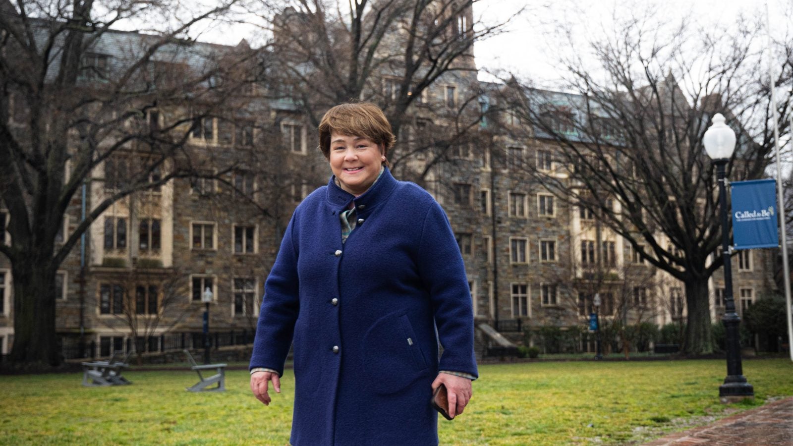 A white woman in a bright blue jacket stands in front of a stone building on Georgetown&#039;s campus smiling.