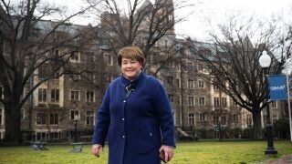 A white woman in a bright blue jacket stands in front of a stone building on Georgetown&#039;s campus smiling.