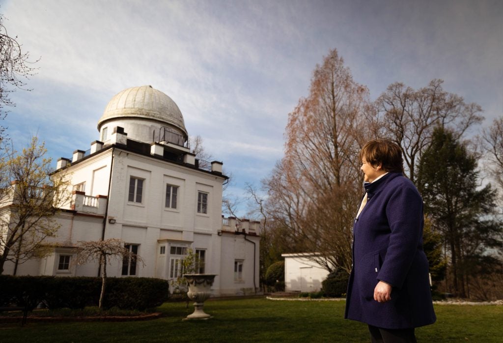 A woman with brown hair and a bright blue jacket looks at the rounded dome of an observatory behind her.
