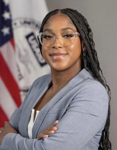 Young Black woman with glasses in formal attire in front of a US flag