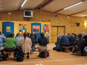 A group of pilgrims on benches talking with a brother at Taize in a yellow room.