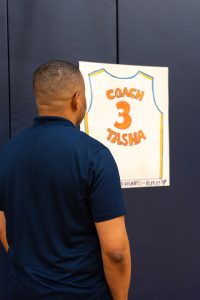 A black man in a blue shirt stares at a poster hanging in a gym that says "Coach Tasha" with the number 3.
