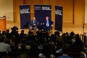 Two men sit on stage discussing politics in front of a crowd of students.