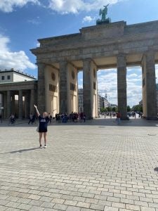 A white woman stands in front of the Brandenburg Gate in Berlin with her hands in the air on a sunny day.