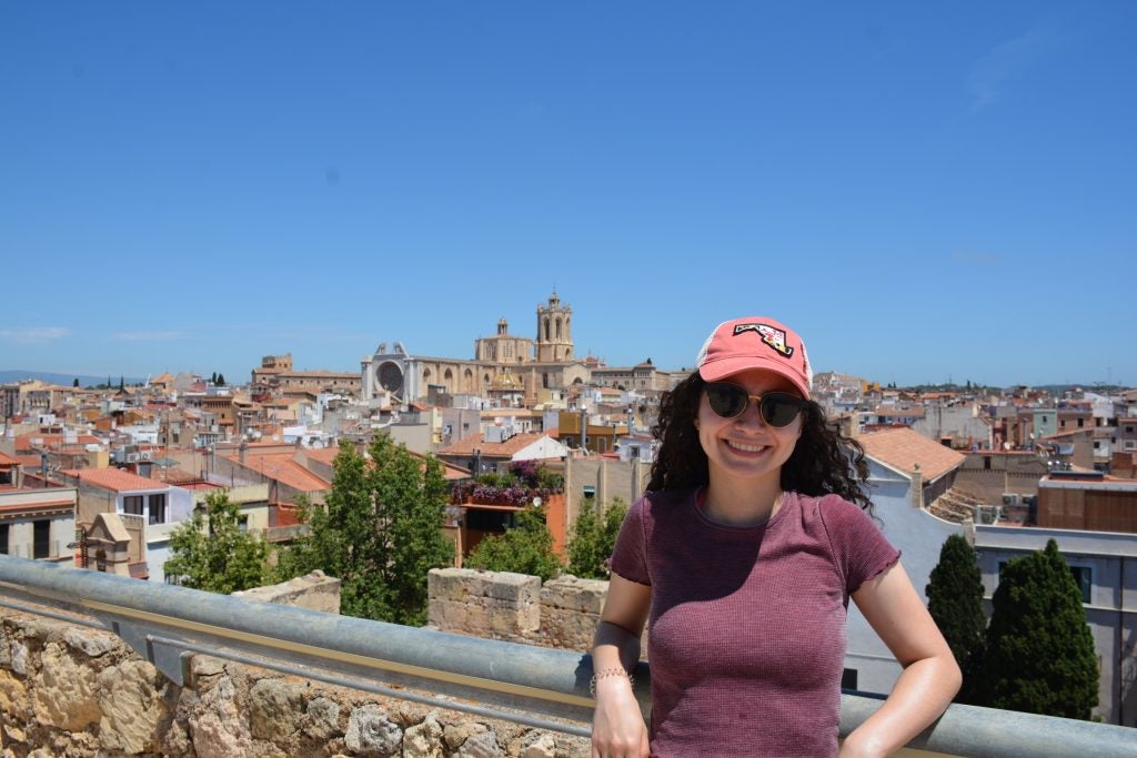 A Colombian woman stands on a patio overlooking a skyline in Spain. She wears a hat and sunglasses.