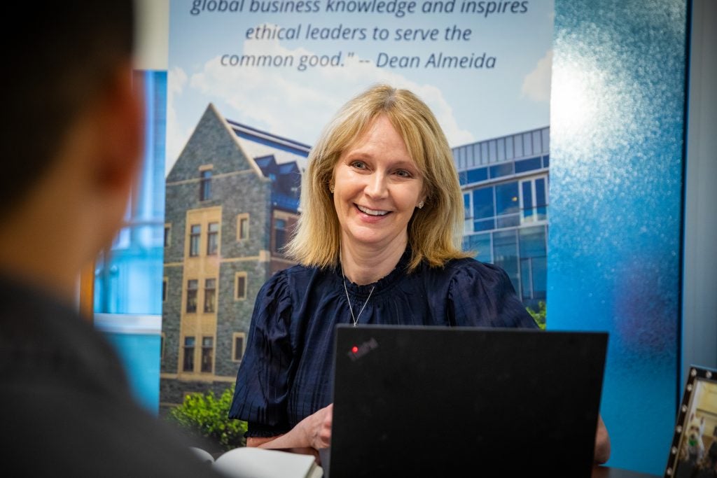 A white woman with blonde hair sits in front of a poster for the McDonough School of Business with a laptop in front of her.
