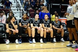 A group of women's basketball players sit on the bench wearing black jerseys.
