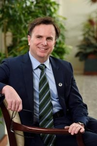 A white man sits on a chair in a suit and tie and smiles.