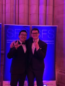 Two Georgetown students in black suits hold up their hands to form their initials in front of a pink curtain and a blue sign. 