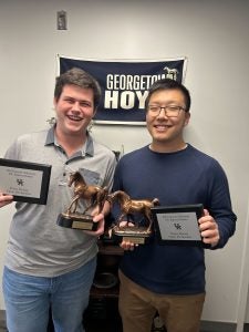 Two male students smile and hold trophies of horses and a plaque. A banner behind them reads "Georgetown Hoyas."