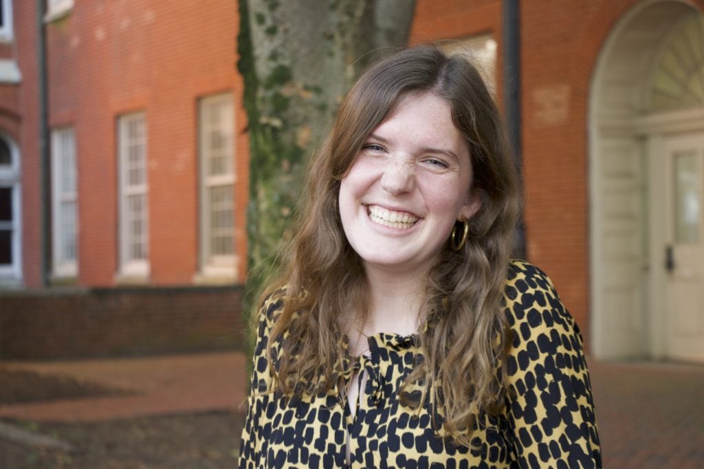 A white woman with brown hair and a patterned shirt smiles in front of a brick building outside.