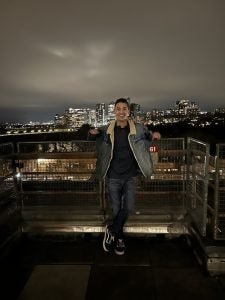 A high school student stands on a balcony overlooking a city at night.