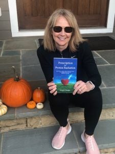 A woman with sunglasses sits on the front stoop of a house holding a book. Next to her are pumpkins.