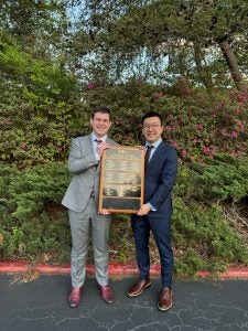 Two male students smile and hold a plaque outside.