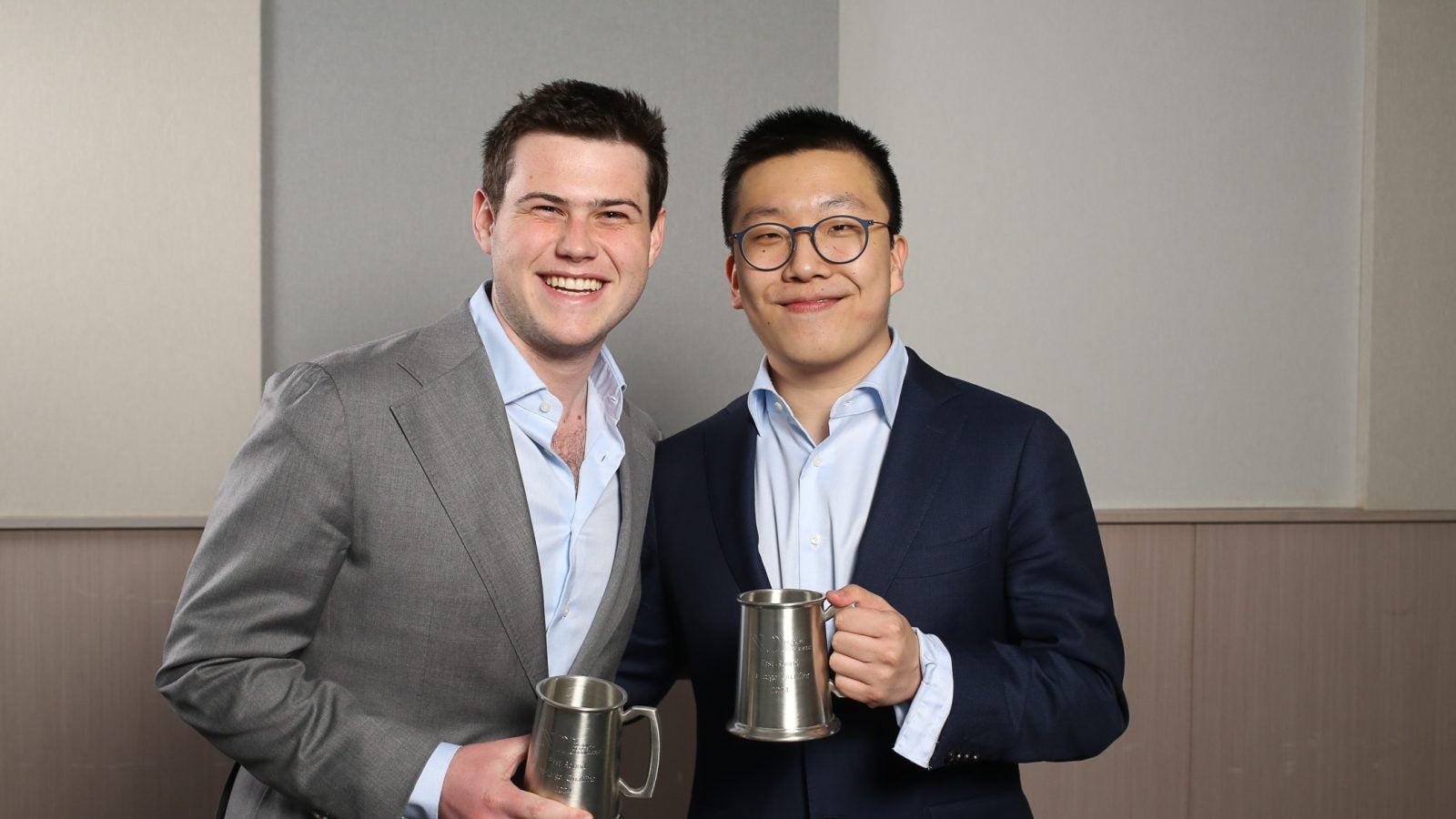 Two male Georgetown students dressed in suits hold metal cups, a prize for a debate tournament.