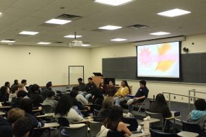 A group of students sit in front of a classroom on a panel while other classmates sit in desks watching them.