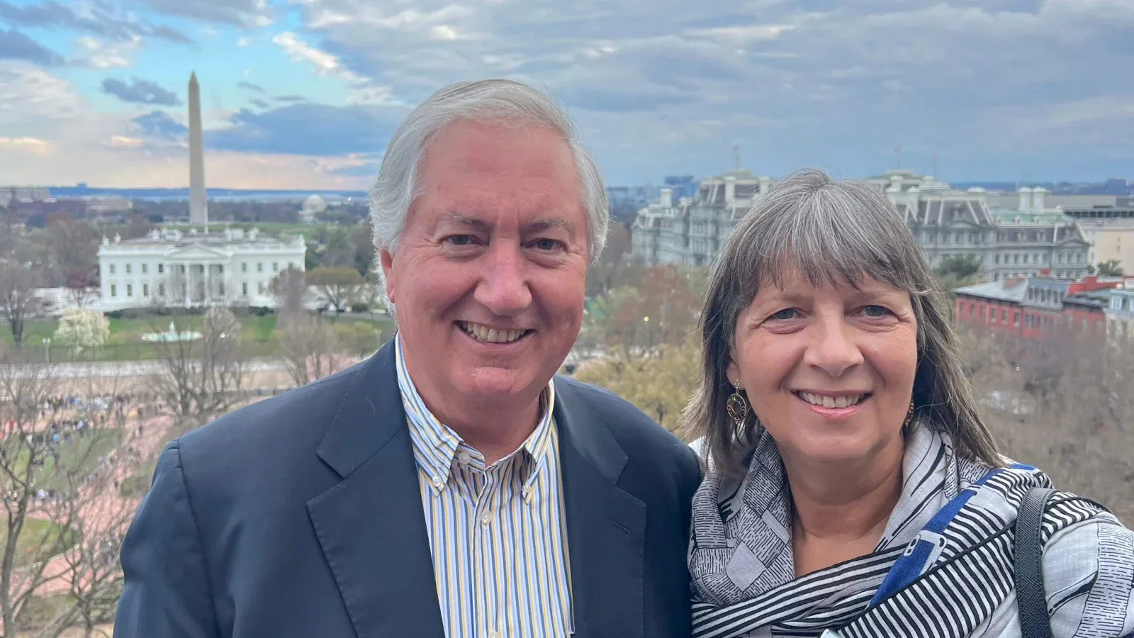 A white man and woman smile together in front of a cloudy sky and buildings.