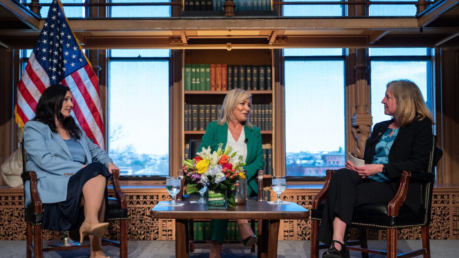 Three women sit on a panel talking with an American flag and a bookcase behind them.