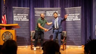 One man waves to a crowd on stage while shaking the hand of the man standing next to him.