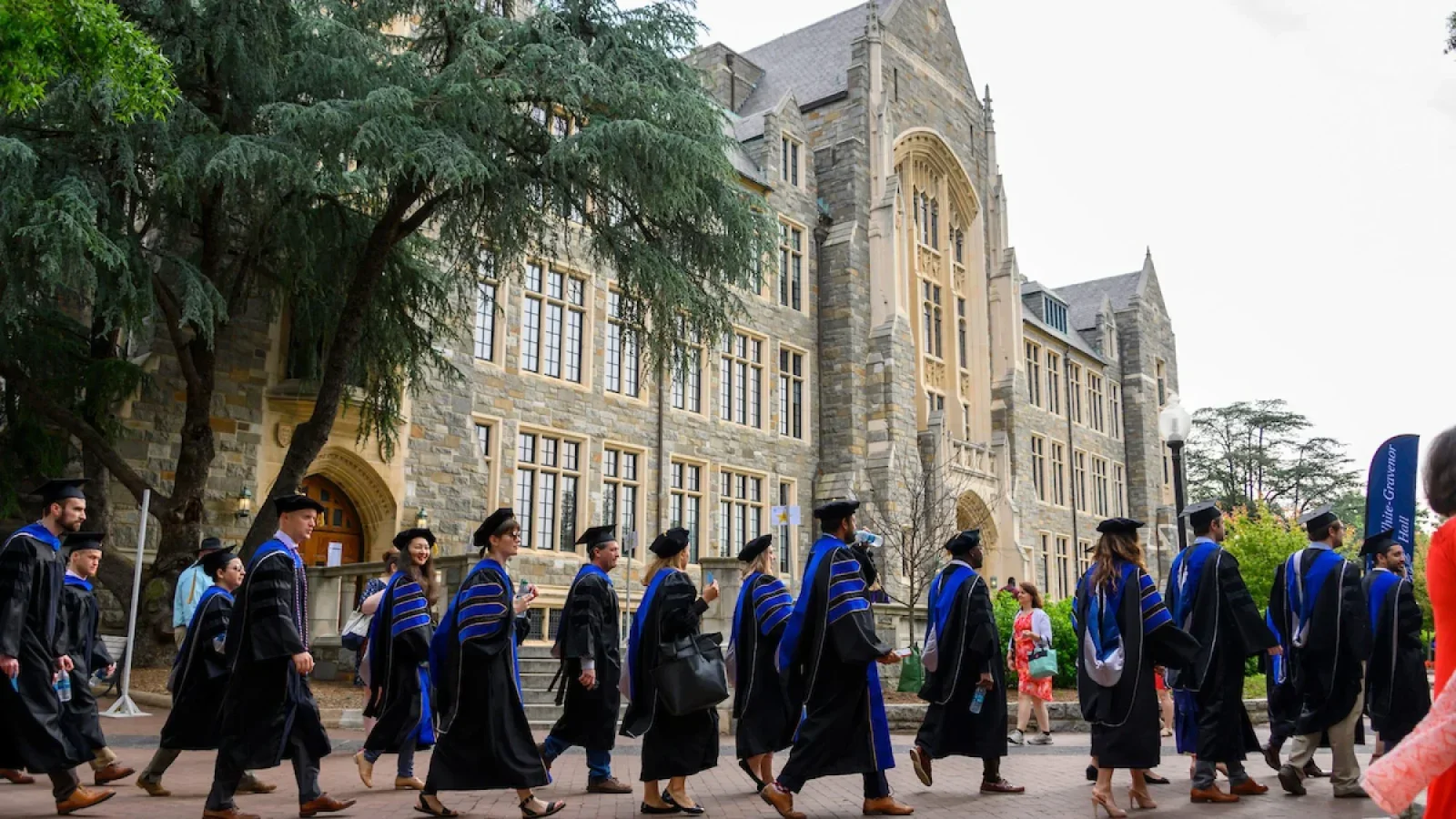 Students in academic robes walk past a stone building.