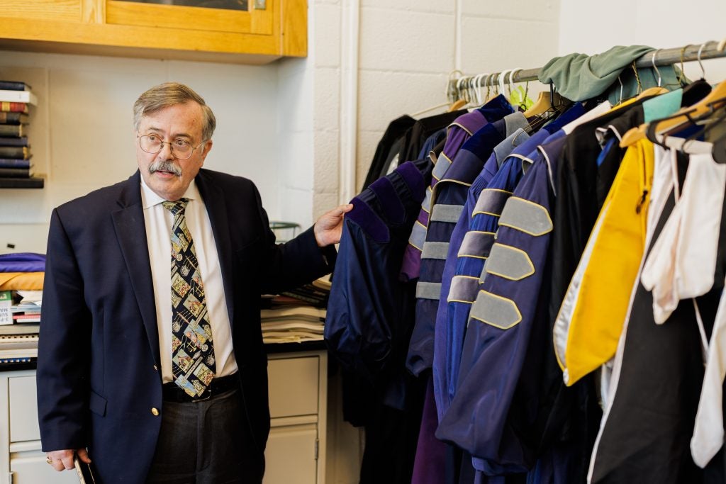 A man with glasses and a mustache wearing a suit and tie holds the sleeve of an academic robes that is next to other robes.