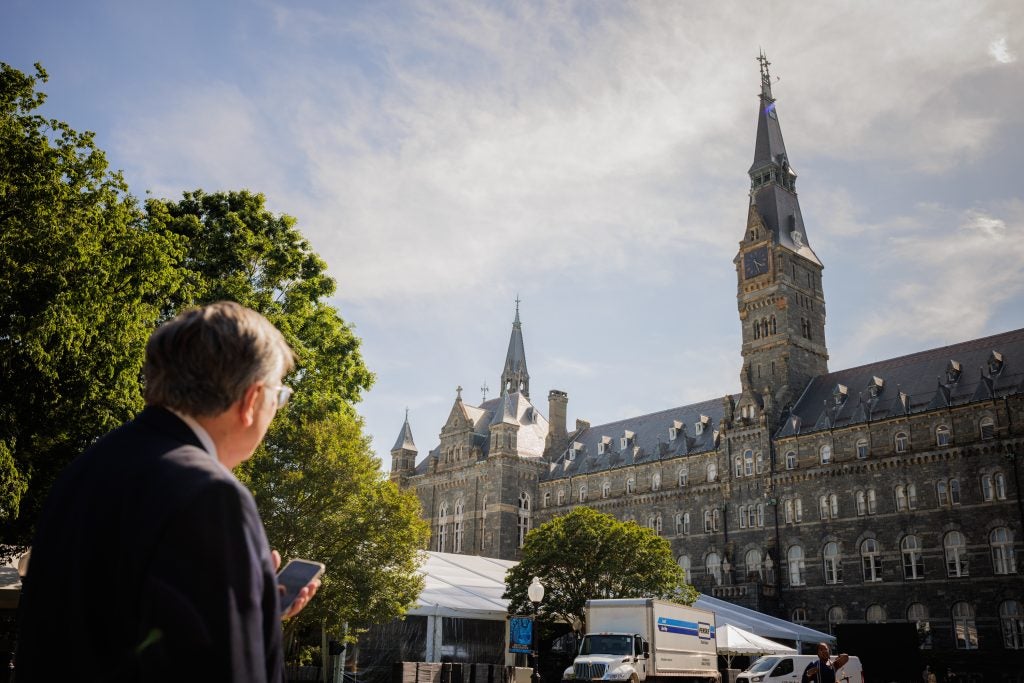 A white man with glasses holds his phone and looks up at a stone building's clock.