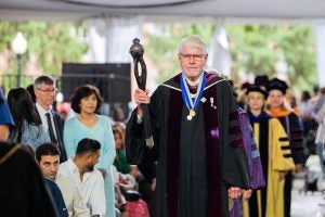 A white man with gray hair and a beard and glasses holds a mace and wears a graduation gown as he processes down an aisle under a tent leading a line of faculty and graduates.