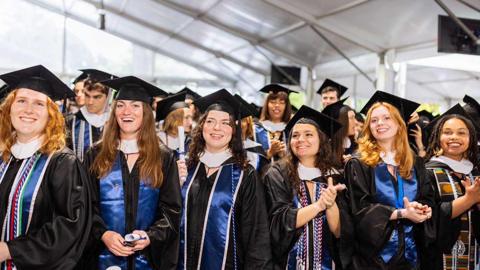 A group of graduates in caps and gowns smile at a commencement ceremony.