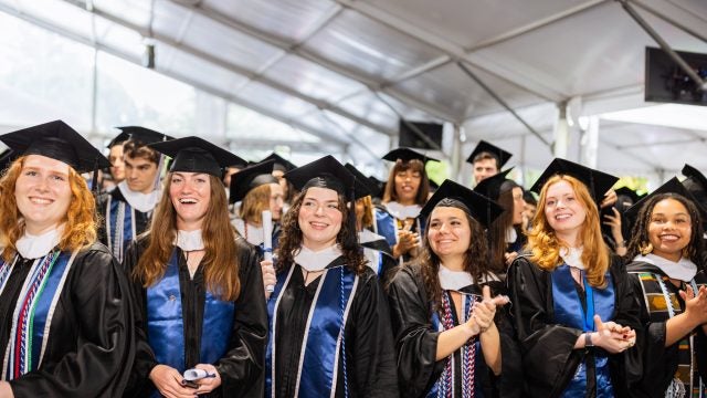 A group of graduates in caps and gowns smile at a commencement ceremony.