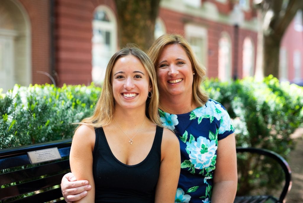 A mother and daughter sit side-by-side on a bench outdoors and smile.