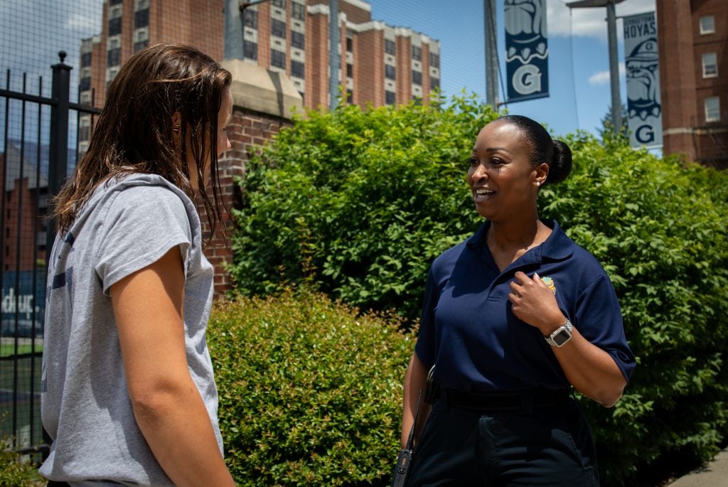 A Black female police officer wearing a navy shirt smiles as she talks with a student in front of Georgetown's athletic field.