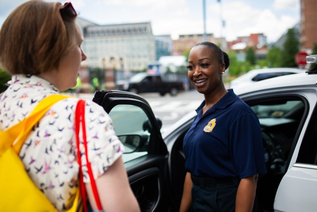 A Black female police officer stands outside her police car and smiles at a student as they talk.