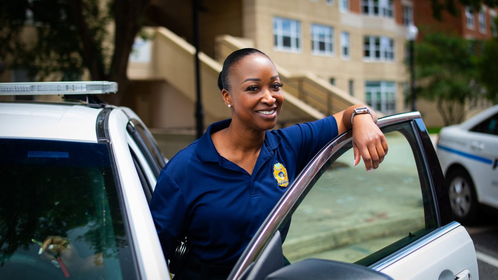 A Black female police officer leans over the door of her police car and smiles.