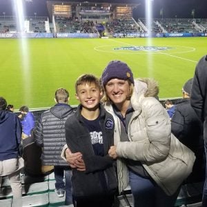 A white woman in a white jacket and blue hat poses with her son in a jacket in front of a soccer field at night.
