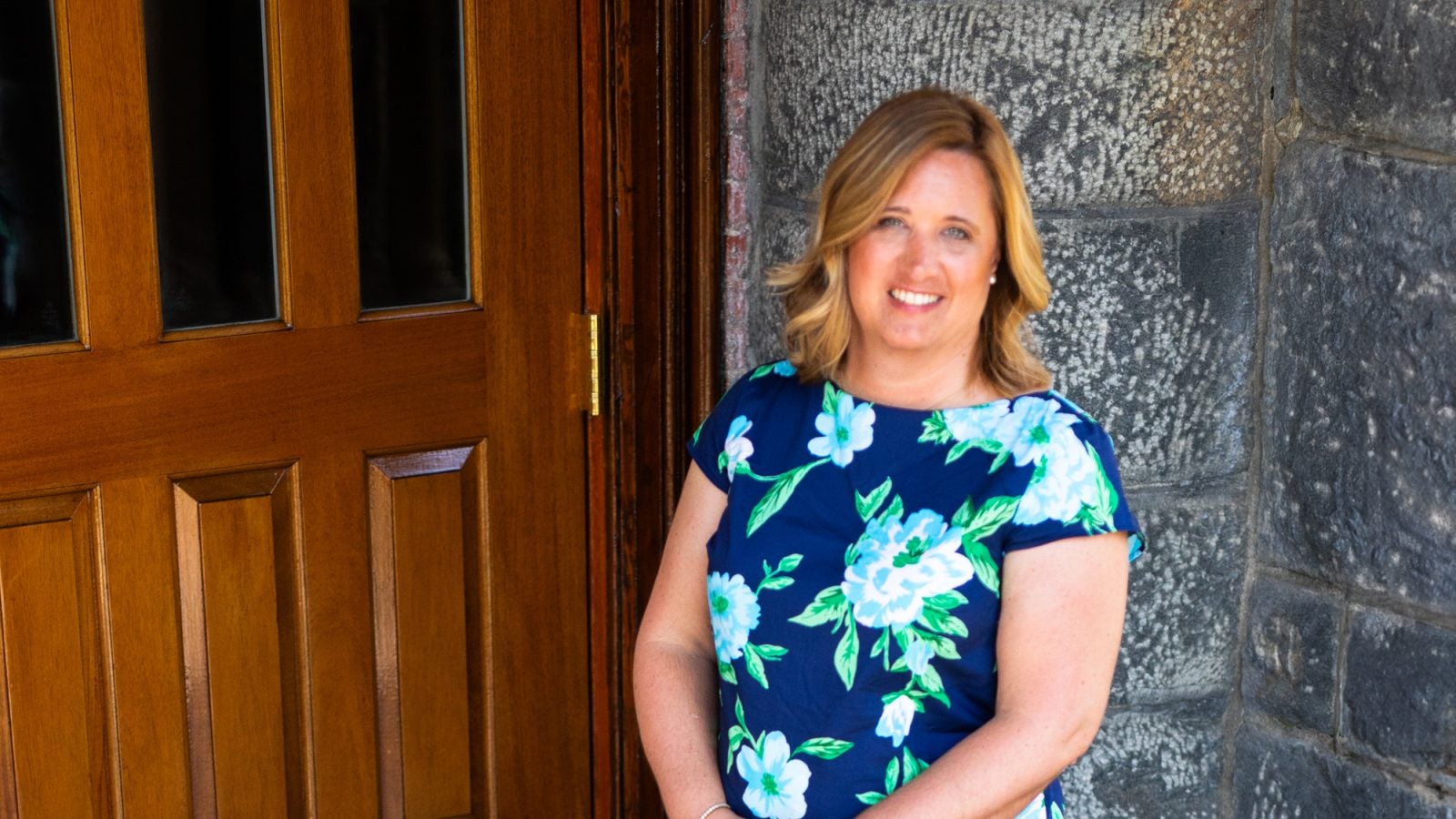 A woman in a floral blue shirt smiles outside the entrance to a building