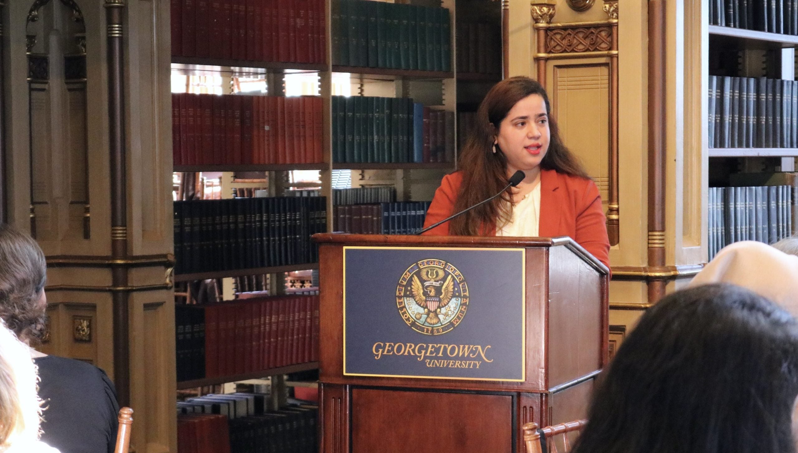 An Afghan woman in an orange blazer speaks behind a podium in a library.