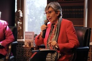 An Afghan woman in a bright pink blazer and scarf holds a microphone and speaks.