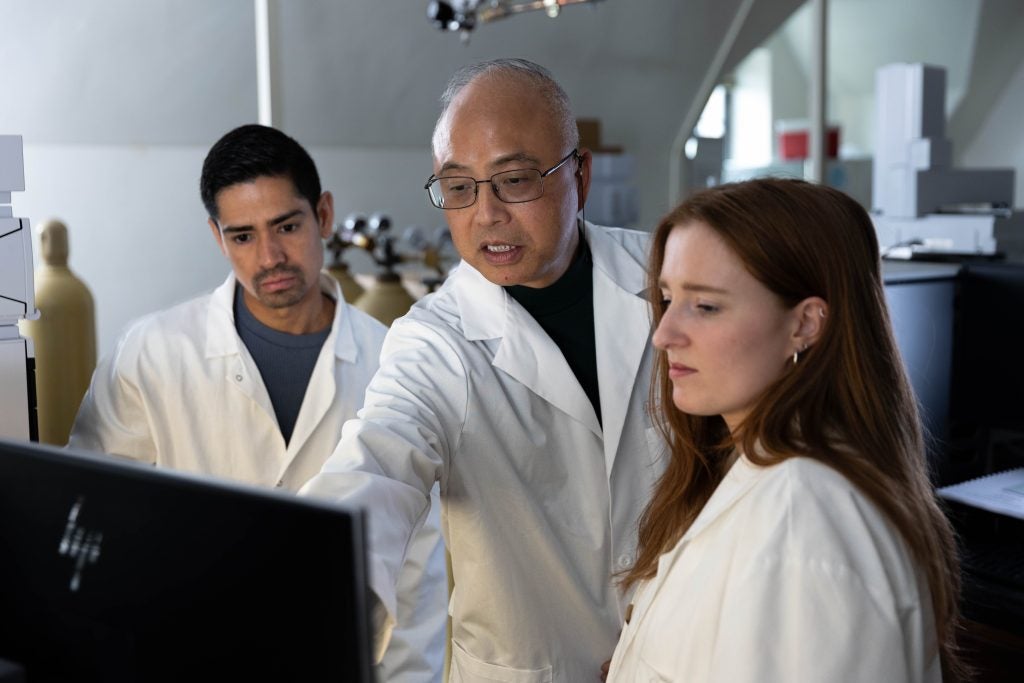 A man wearing a white lab coat and glasses points to a computer in the middle of two students in a laboratory.