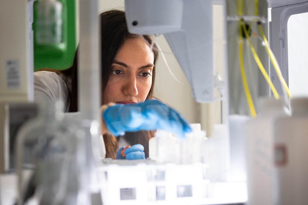 A student wearing blue gloves and a white lab coat works on a machine in a lab.