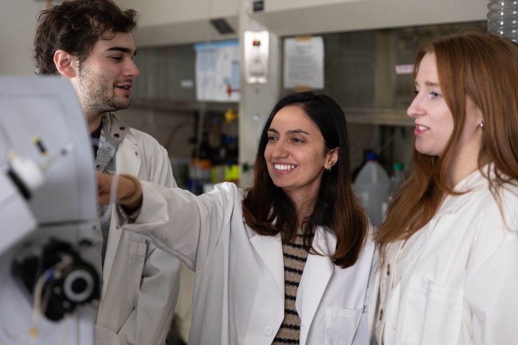 Three students are in a lab wearing white lab coats.
