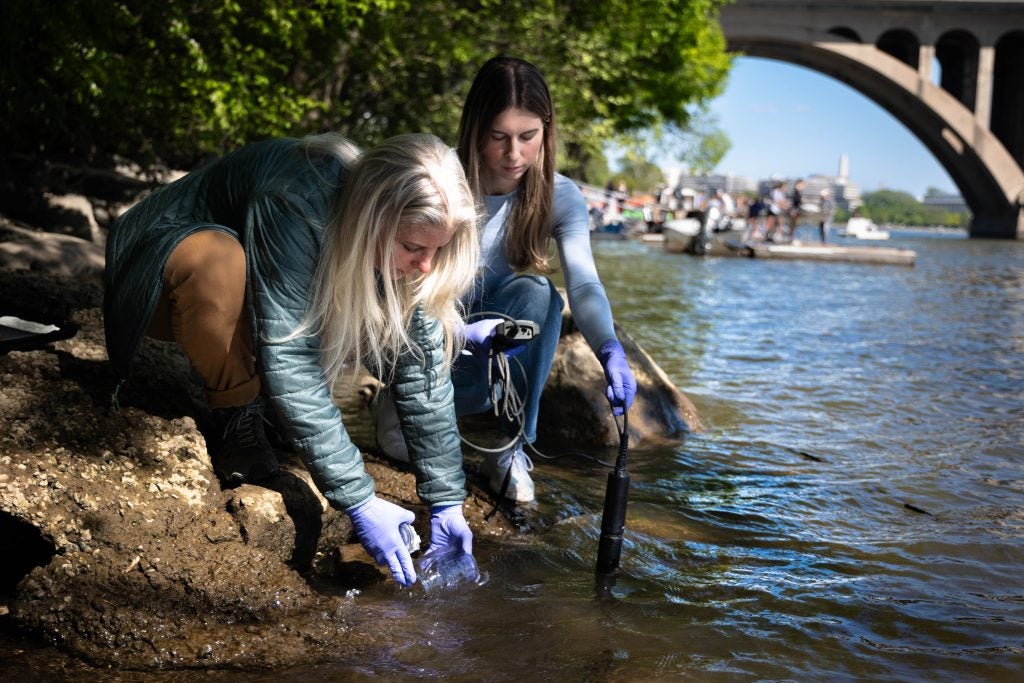 A woman dips a jar into the Potomac River wearing purple gloves next to a student who measures the water on a sunny day.