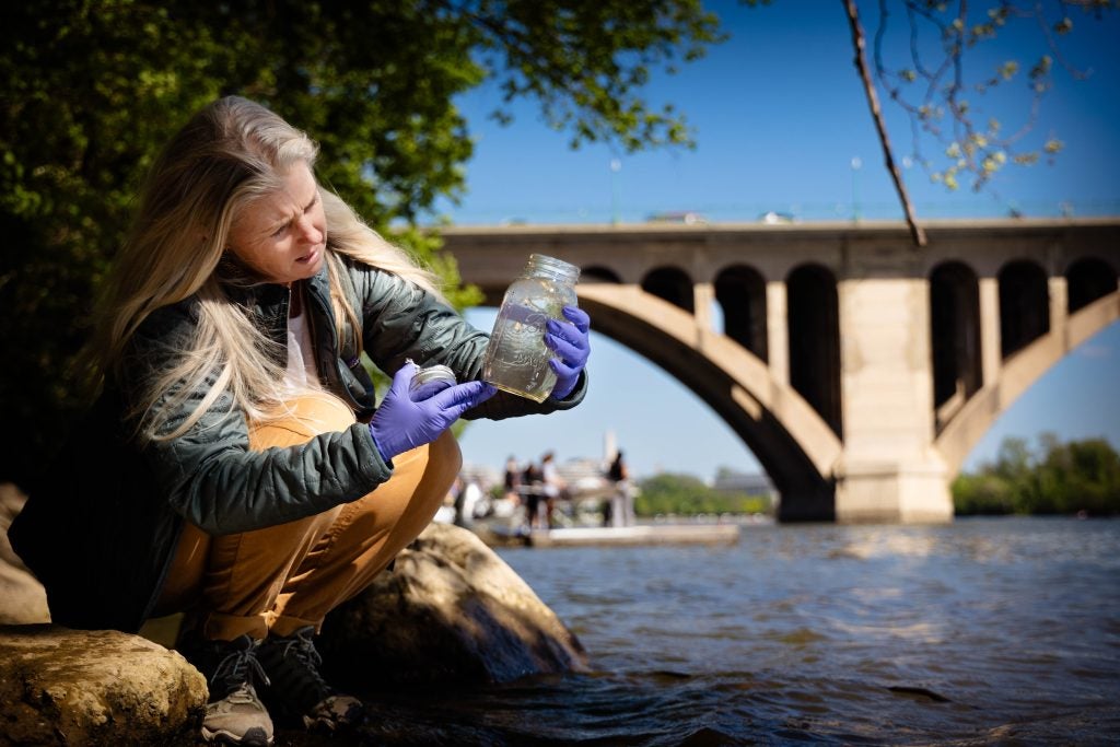 A woman holds up a jar of water on the shore of a river with a bridge behind her.