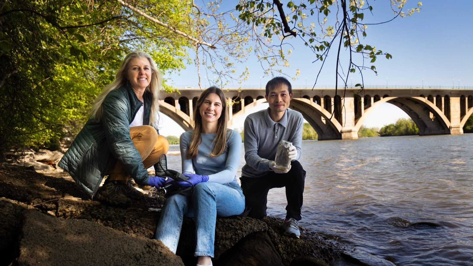 A student and two professors pose on the rocky shoreline of the Potomac River in front of a bridge on a sunny day.