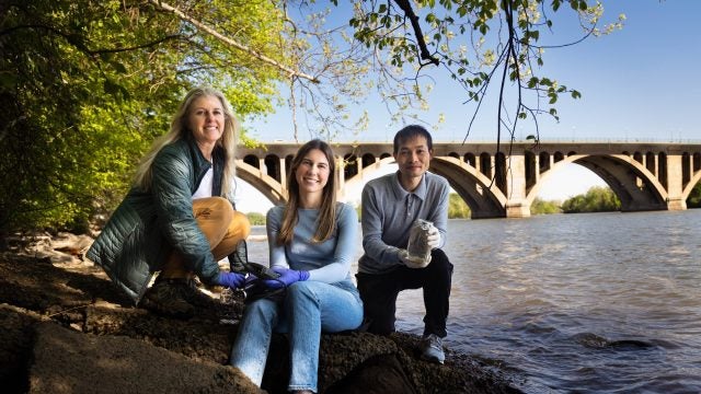 A student and two professors pose on the rocky shoreline of the Potomac River in front of a bridge on a sunny day.