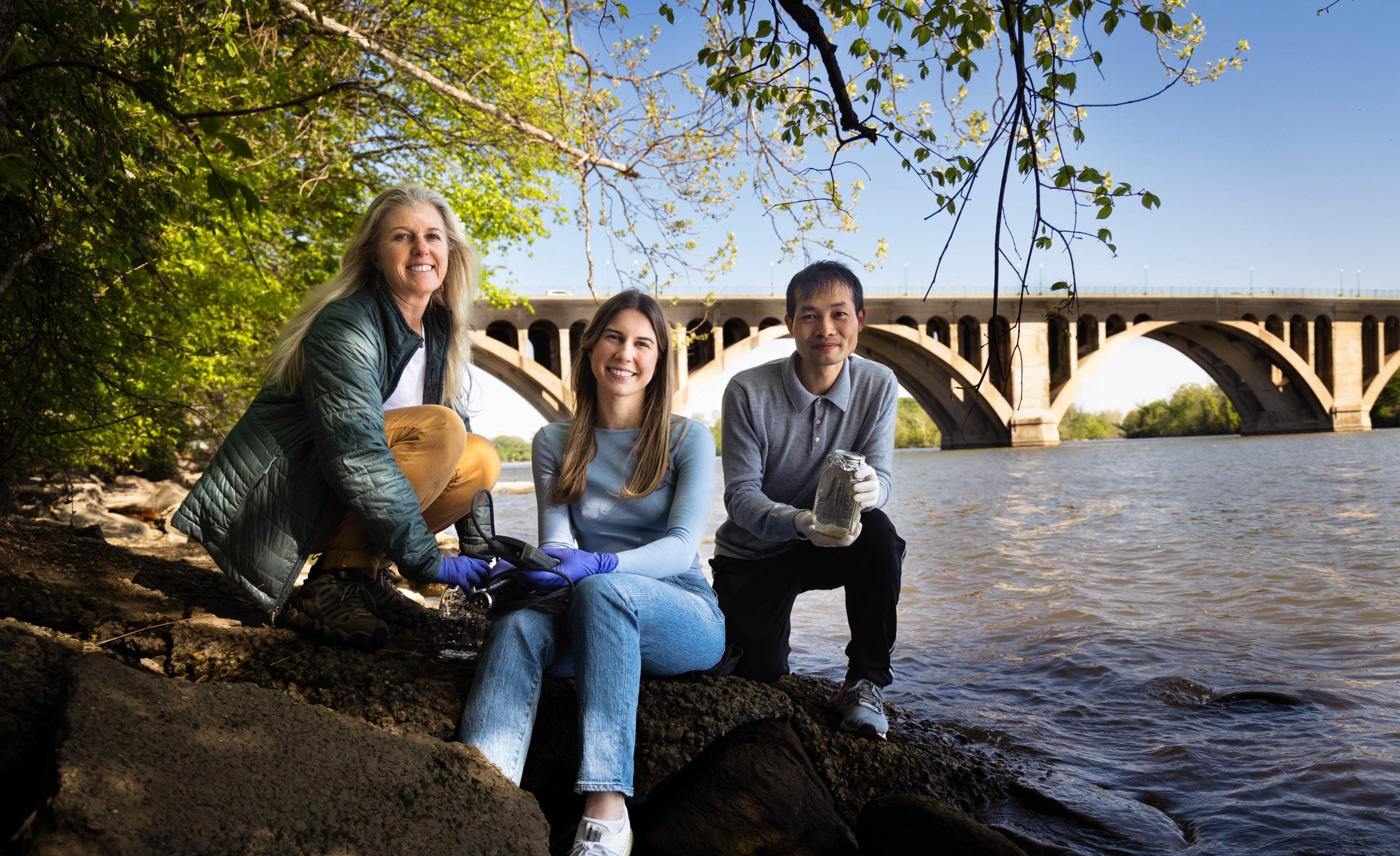 A student and two professors pose on the rocky shoreline of the Potomac River in front of a bridge on a sunny day.